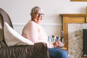 Portrait of happy laughing senior woman using phone while sitting on armchair at home. Mature older
