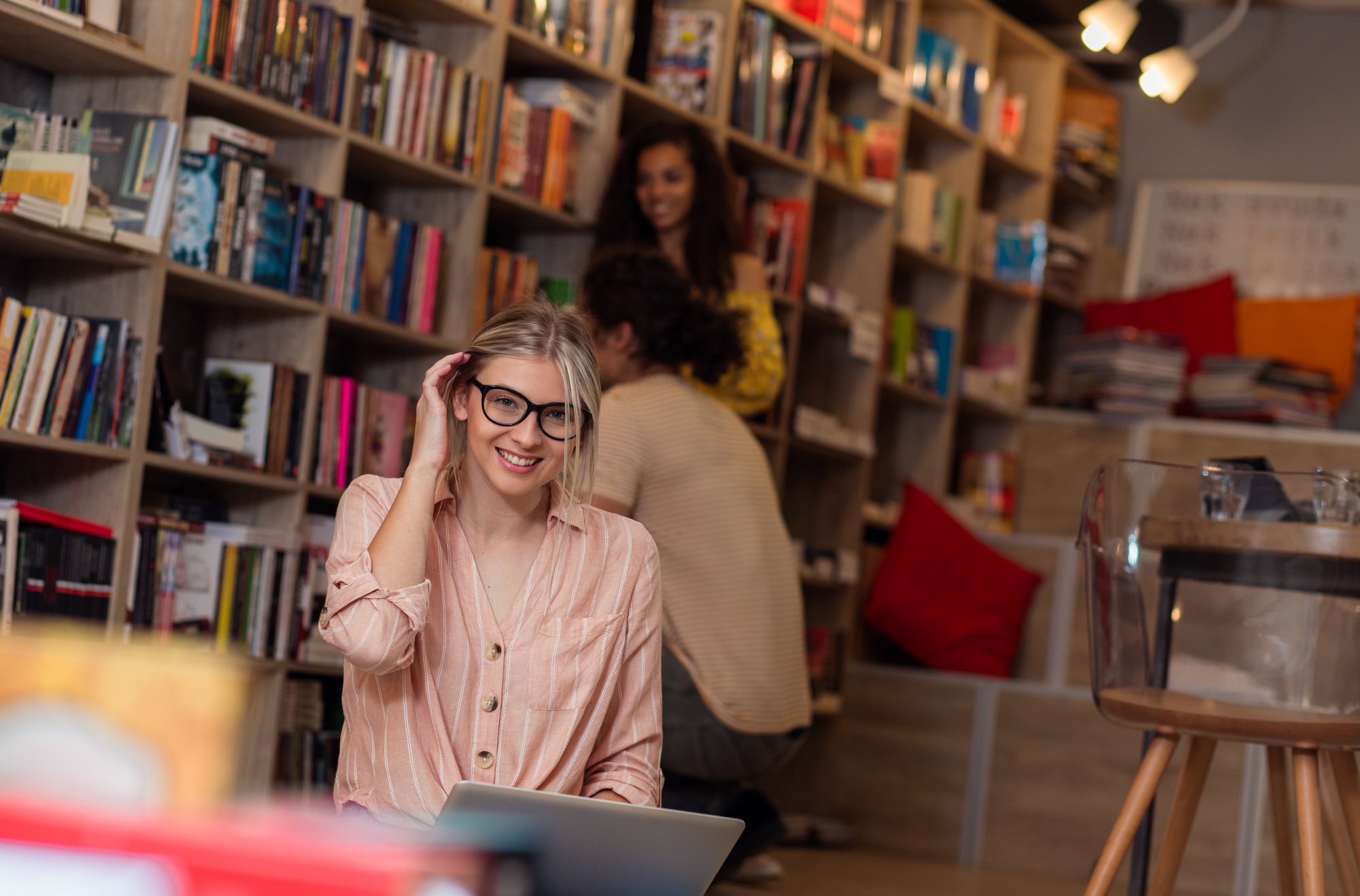 Portrait of young female student sitting at library using laptop with her friends in background.